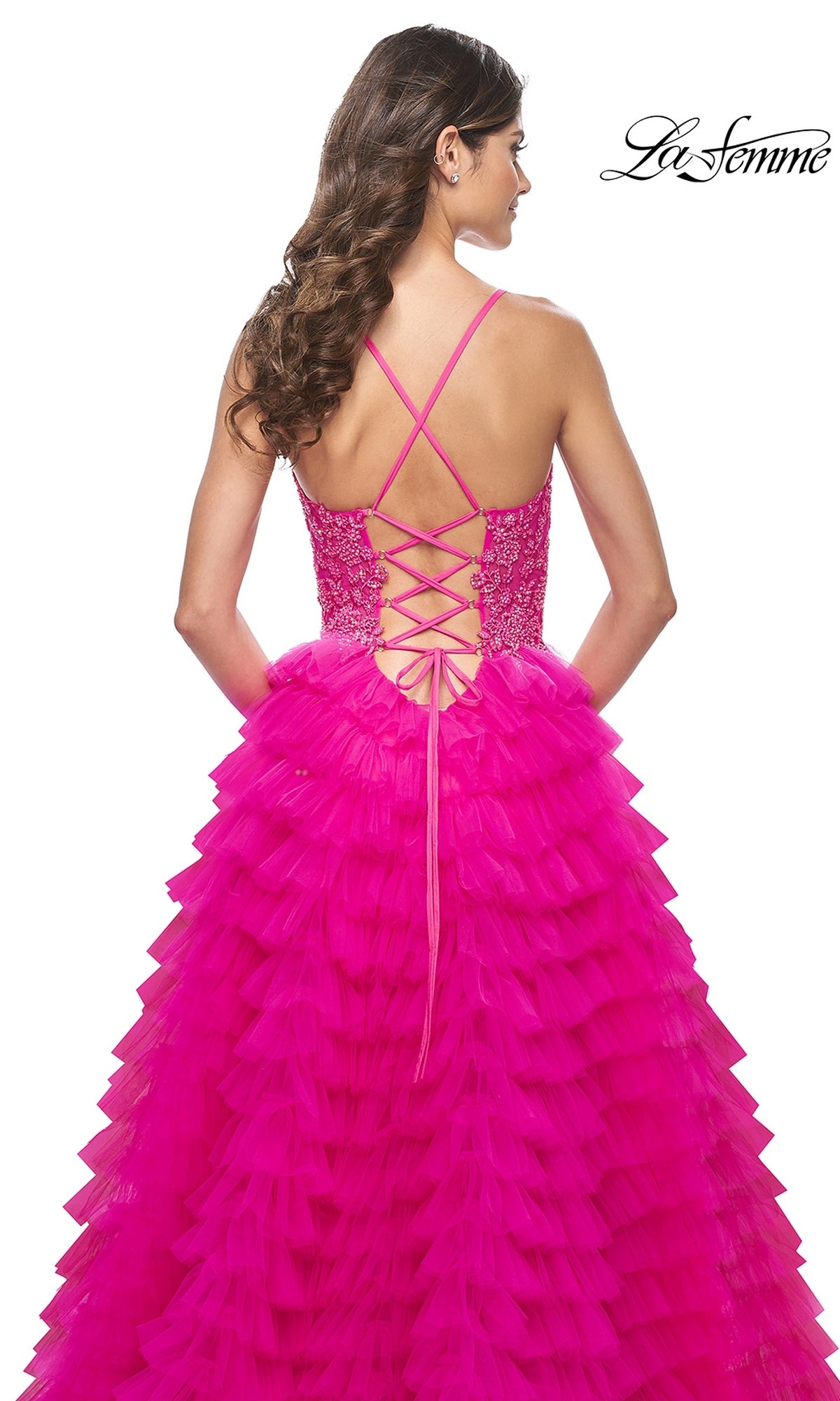 A woman models the La Femme Hot Pink Long Ruffled Prom Dress 32334, featuring an embroidered lace bodice, ruffled A-line skirt, and lace-up back, shown from behind against a white background.